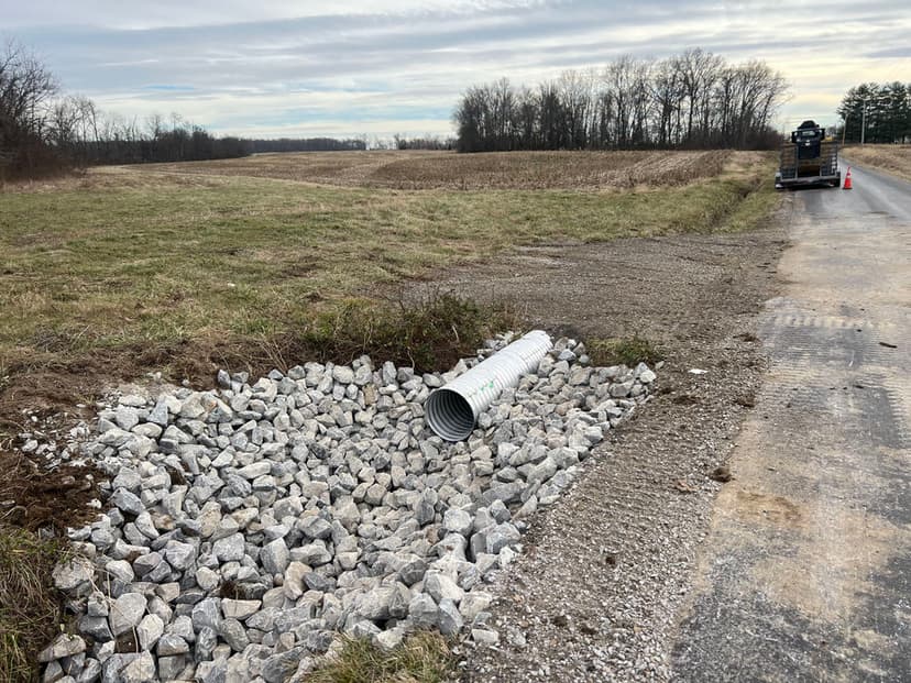 Drainage pipe installation with gravel at road edge in rural landscape.