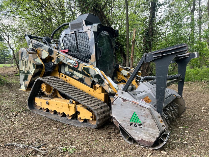 Camouflaged track-mounted skid steer loader with mulching attachment in a wooded area.